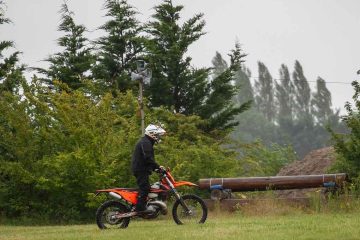 Homme sur motocyclette orange dans un champ herbeux, entouré d'arbres, sous un ciel gris