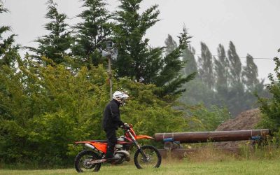 Homme sur motocyclette orange dans un champ herbeux, entouré d'arbres, sous un ciel gris